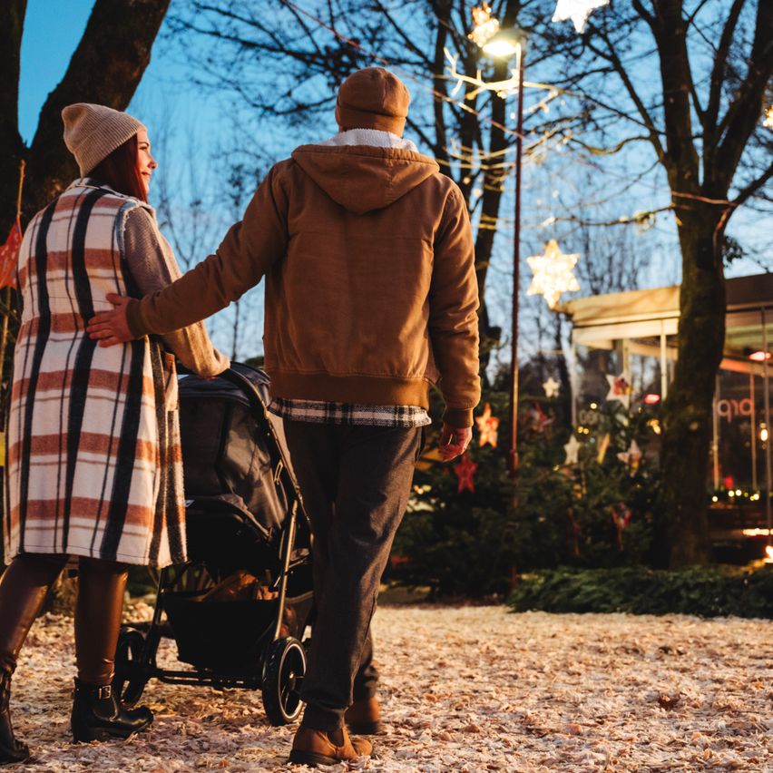 Ein Paar geht Hand in Hand mit einem Kinderwagen in einem festlich dekorierten Außenbereich, geschmückt mit Lichtern und Bäumen.