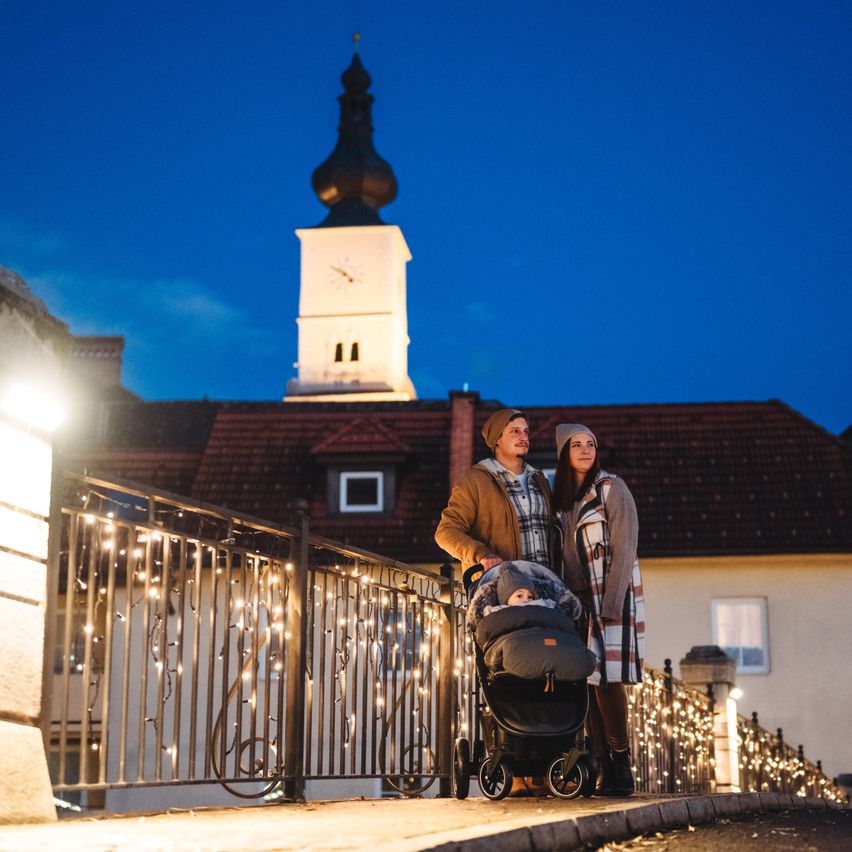 Ein Paar mit einem Baby im Kinderwagen steht auf einer Brücke vor einem Haus mit Turm und Weihnachtsbeleuchtung.