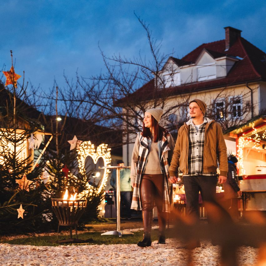 Ein Paar geht bei Nacht auf einem Weihnachtsmarkt Hand in Hand. Sie sind von beleuchteten Bäumen und festlichen Dekorationen umgeben.