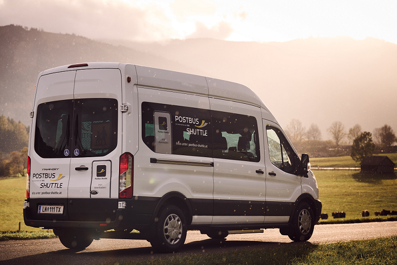 Ein weißer Postbus Shuttle-Van steht am Straßenrand, mit einer Berglandschaft im Hintergrund.