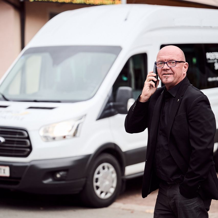 Ein kahlköpfiger Mann in einem schwarzen Anzug und mit Brille geht und spricht auf seinem Handy vor einem weißen Lieferwagen.
