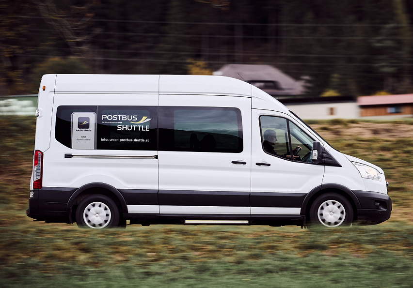 Ein weißer Postbus Shuttle-Van fährt auf einem Grasfeld. Der Van hat einen schwarzen Rand und trägt den Text 'POSTBUS SHUTTLE' auf seiner Seite.