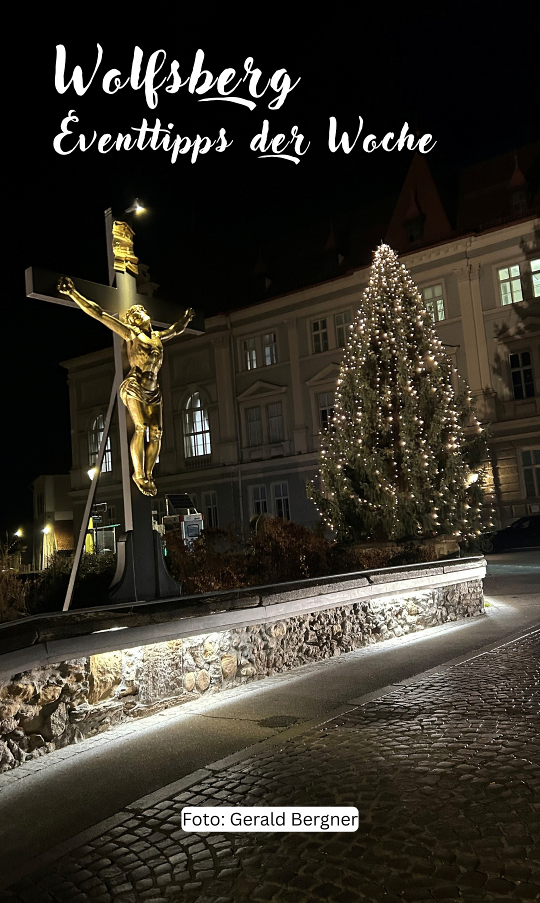 Ein goldenes Kruzifix steht beleuchtet in der Nacht neben einem geschmückten Weihnachtsbaum vor einem Gebäude mit vielen Fenstern.