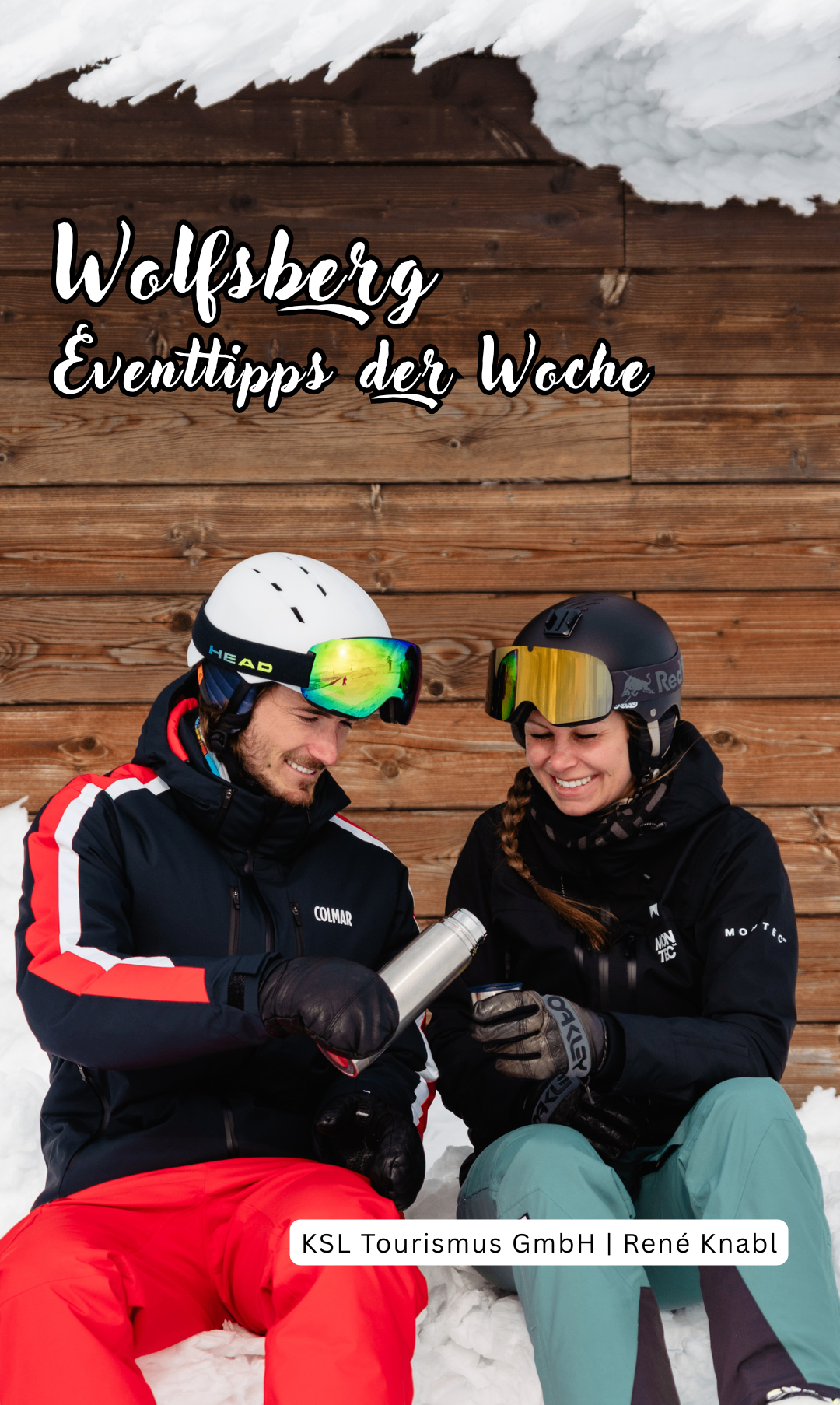 Zwei Skifahrer, ein Mann und eine Frau, sitzen im Schnee und tragen Winterkleidung. Sie haben Helme und Schutzbrillen. Hinter ihnen ist eine Holzwand.