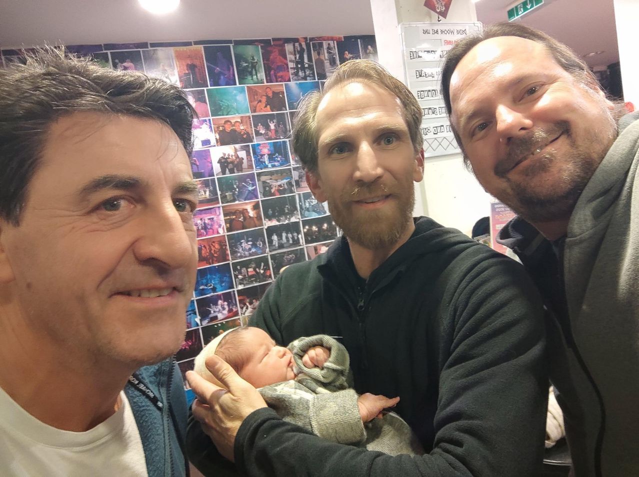 Drei Männer lächeln und posieren für ein Foto mit einem Neugeborenen in einem Raum mit an der Wand montierten Bildern.