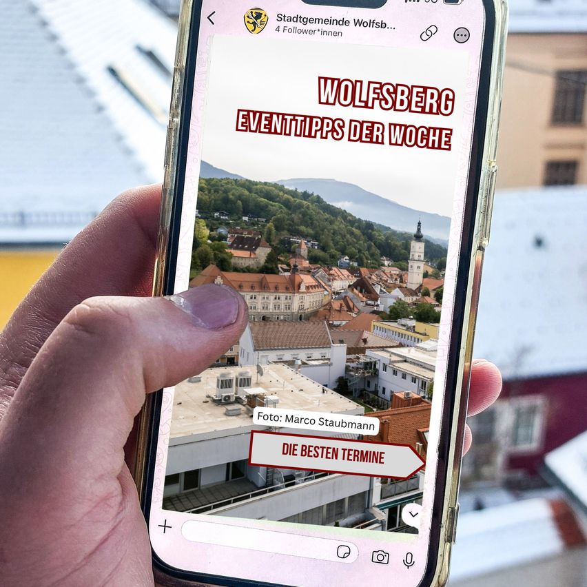 Eine Hand hält ein Telefon, das eine Stadtansicht mit Wolfsberg Eventtipps der Woche in rotem Text anzeigt. Das Foto ist von Marco Staubmann.