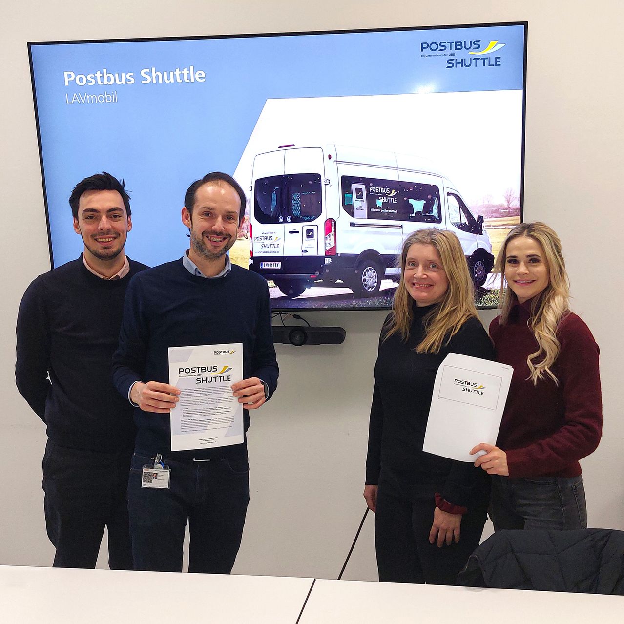 Vier Personen stehen lächelnd vor einer großen Leinwand. Die Leinwand zeigt einen weißen Van mit der Aufschrift 'Postbus Shuttle LAVmobil'. Jede Person hält ein Zertifikat mit demselben Text.