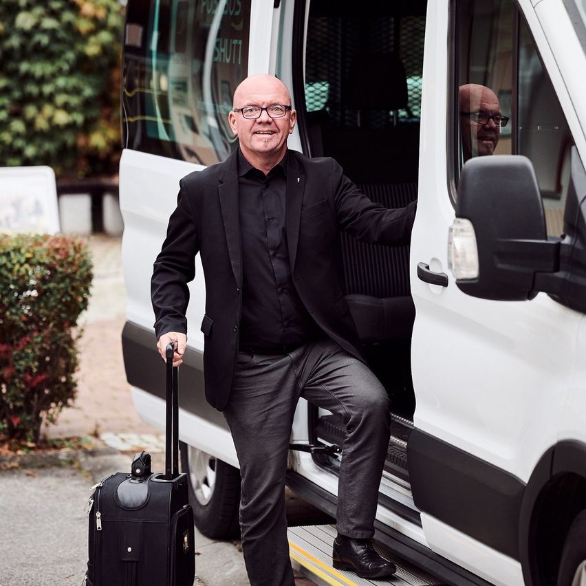 Ein Mann steigt mit einem Koffertrolley in der Hand aus einem weißen Van aus. Er lächelt und ist in einem schwarzen Anzug gekleidet.