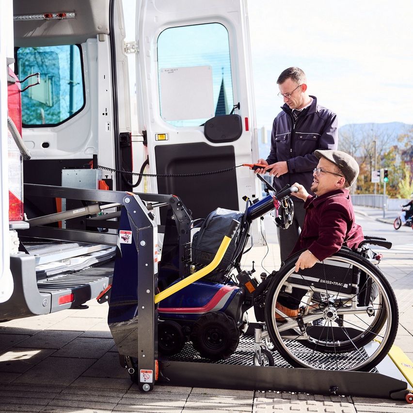 Ein Mann steht und hält eine Fernbedienung, während ein anderer Mann im Rollstuhl dabei geholfen wird, in ein Fahrzeug einzusteigen.