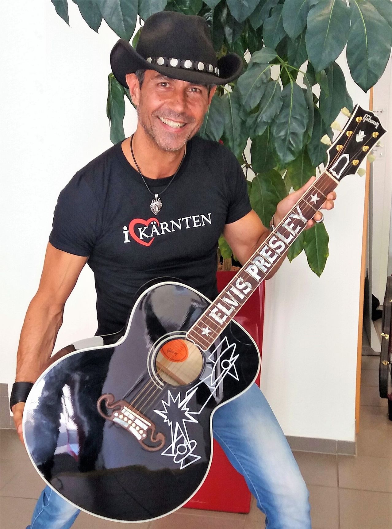 Ein Mann hält eine Elvis Presley Gitarre und lächelt, mit einem Shirt, das 'i Kärnten' sagt, und einem schwarzen Cowboyhut. Er sitzt mit einer Pflanze hinter ihm.