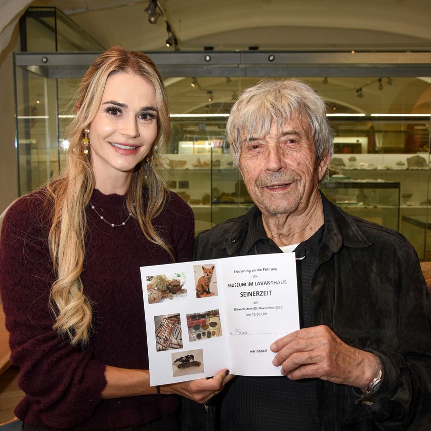 Ein älterer Mann und eine junge Frau stehen in einem Museum, lächeln und halten ein Zertifikat. Die Frau trägt einen Weinroten Pullover und Ohrringe. Der Mann trägt einen schwarzen Mantel und hat eine Uhr am linken Handgelenk. Sie schauen beide in die Kamera. Hinter ihnen steht eine Glasvitrine mit verschiedenen Gegenständen.
