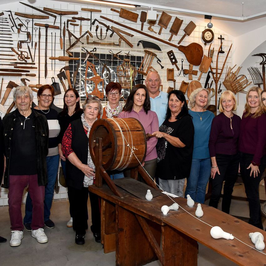 Eine Gruppe von Menschen steht um einen großen Holzfass und Werkzeuge, die an einer Wand montiert sind. Sie scheinen für ein Foto zu posieren, wobei einige das Fass halten.