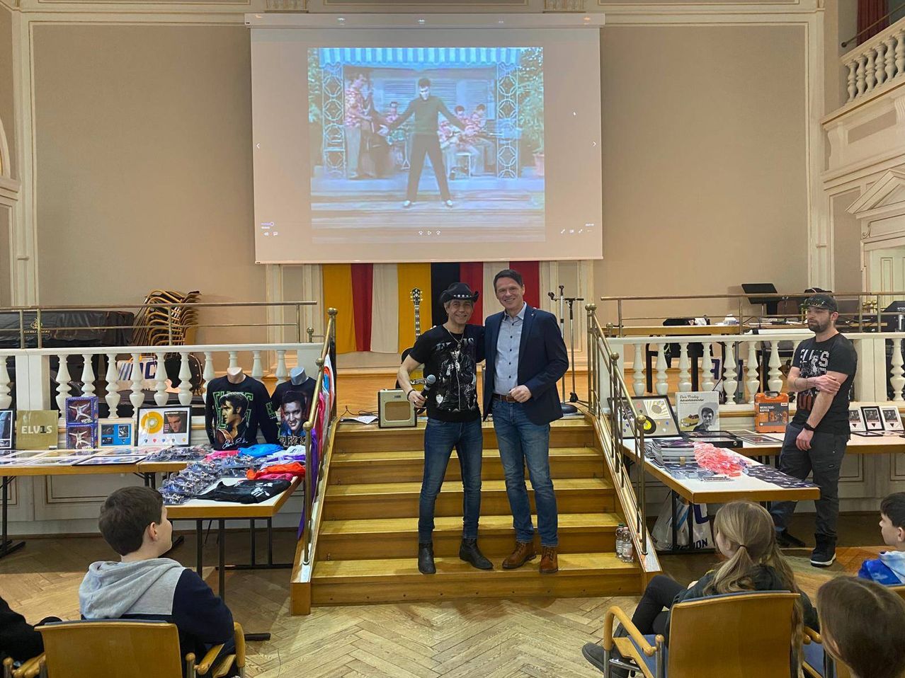 Zwei Männer stehen auf einer Treppe, einer hält ein Mikrofon und der andere trägt einen Cowboyhut. Hinter ihnen befindet sich eine Projektionsleinwand mit einem Film. Links sitzt ein Junge auf einem Stuhl. Rechts sitzt ein Mädchen auf einem Stuhl.