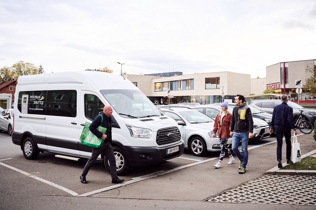 Drei Personen gehen auf einem Parkplatz neben einem weißen Van. Sie werden von einem Mann mit einer grünen Tasche begleitet. Ein weißes Auto ist in der Nähe geparkt. Der Parkplatz ist von Gebäuden umgeben.