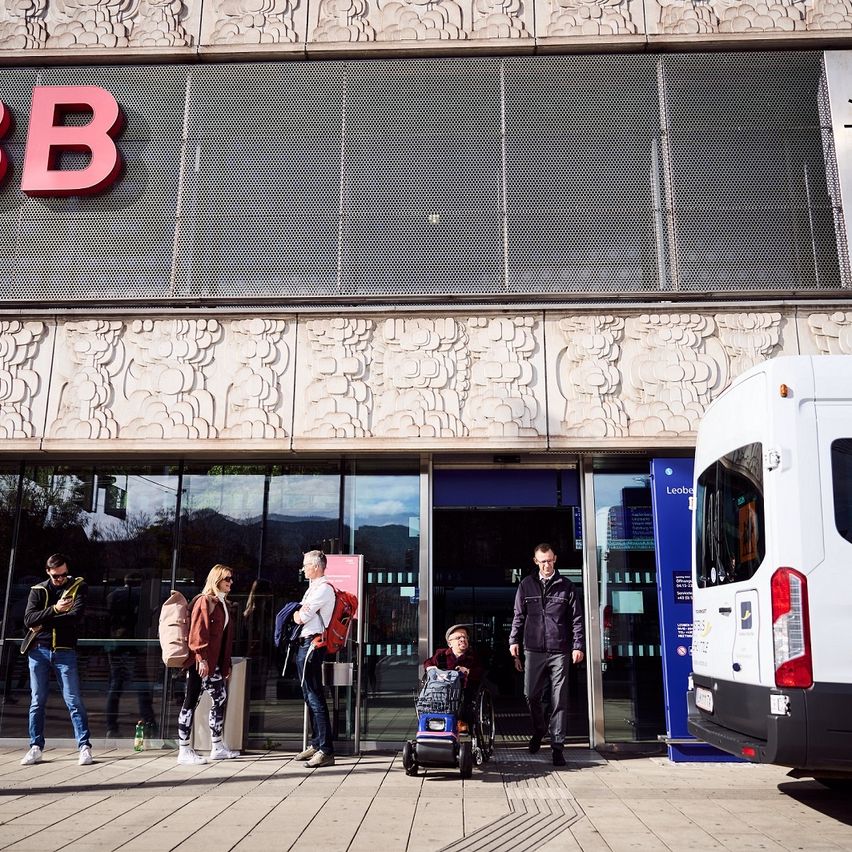 Mehrere Personen stehen vor einem Gebäude mit Glaswänden, möglicherweise einem Bahnhof, mit einem geparkten Van in der Nähe. Das Gebäude hat einen großen roten Buchstaben B an seiner Fassade.