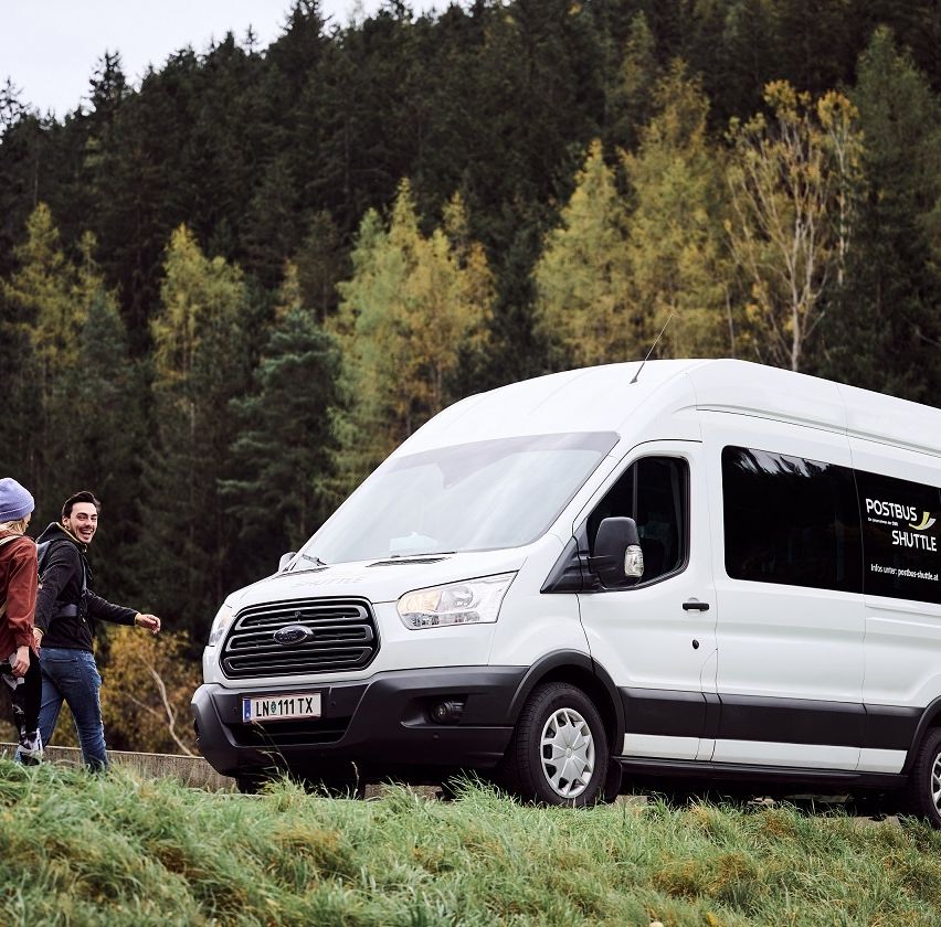Ein weißer Van mit dem Wort POSTBUS an der Seite steht auf dem Gras. Dahinter gehen zwei Personen, eine mit einem Rucksack. Der Hintergrund ist ein Wald mit einer Mischung aus grünen und gelben Blättern.
