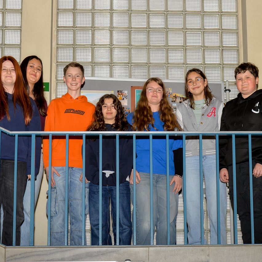 Sieben Teenager stehen auf einem Balkon und lächeln für ein Foto. Sie tragen Freizeitkleidung. Hinter ihnen ist eine Glaswand mit einem weißen Gitter.
