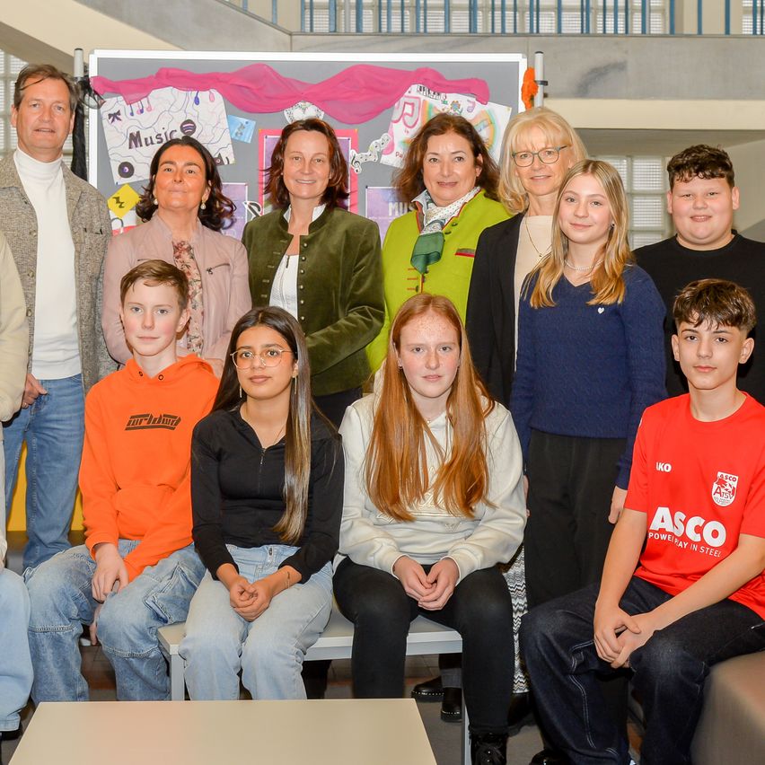 Eine Gruppe von Erwachsenen und Kindern posiert für ein Foto vor einer Wand mit farbenfrohen Dekorationen. Die Erwachsenen stehen, während die Kinder auf einer Bank sitzen.