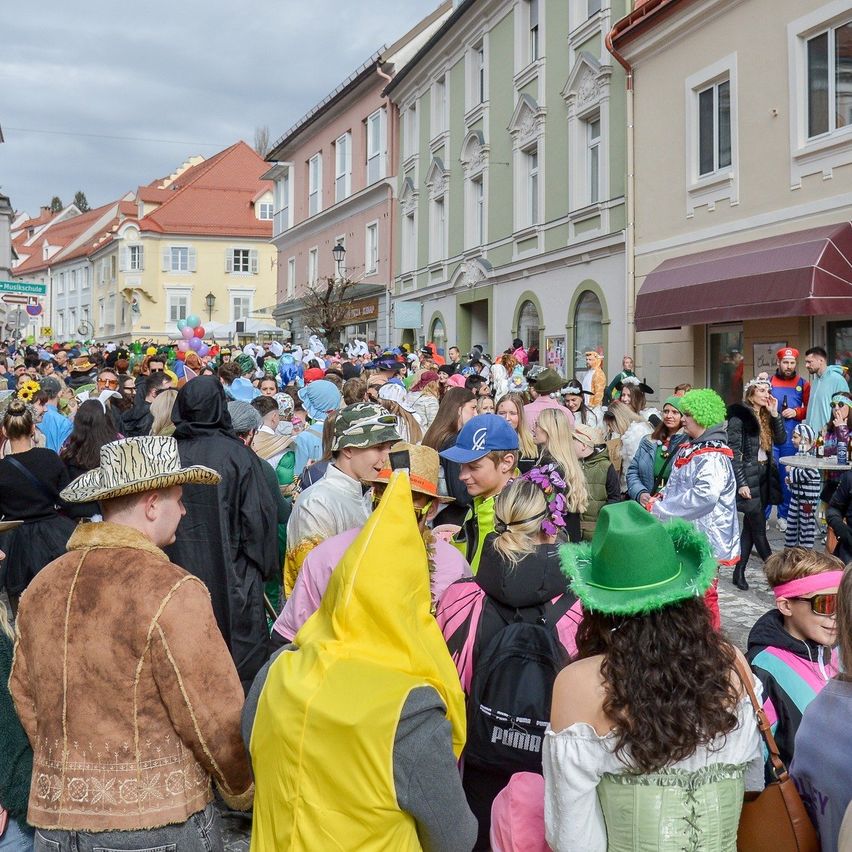 Eine Menschenmenge in Kostümen läuft eine Stadtstraße entlang, einige tragen Hüte und andere sind in vollständigen Kostümen, vor Gebäuden mit Vordächern und Fenstern.