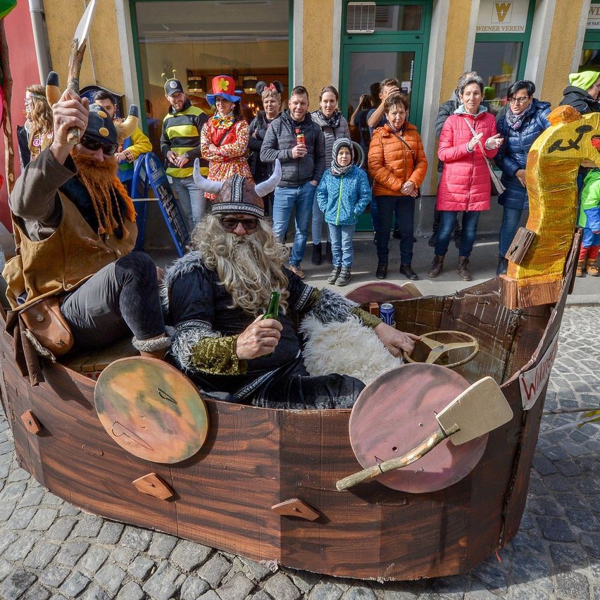 Zwei Personen in Wikingerkostümen sitzen in einem Boot auf einer Kopfsteinpflasterstraße. Dahinter beobachtet eine Menschenmenge in verschiedenen Kostümen.