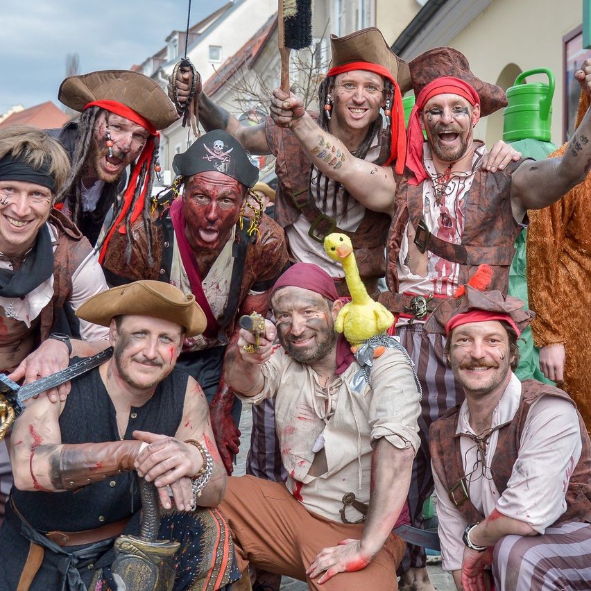 Eine Gruppe von Männern, die als Piraten verkleidet sind, posiert für ein Foto. Sie haben Gesichtsbemalung und halten Requisiten wie Schwerter und ein gelbes Plüschtier.