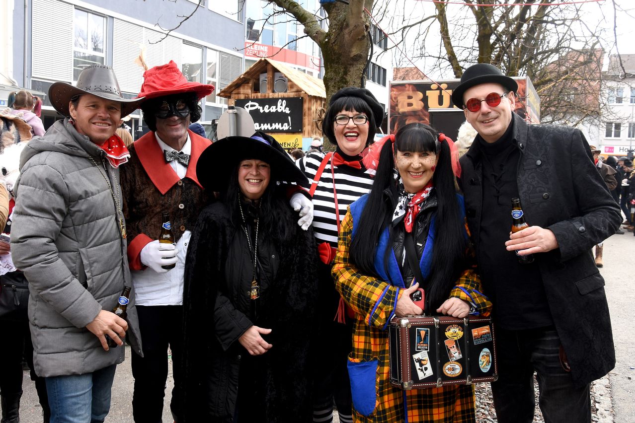 Eine Gruppe von Menschen in Kostümen posiert für ein Foto. Eine Frau mit einem Koffer voller Aufkleber hält eine Bierflasche.