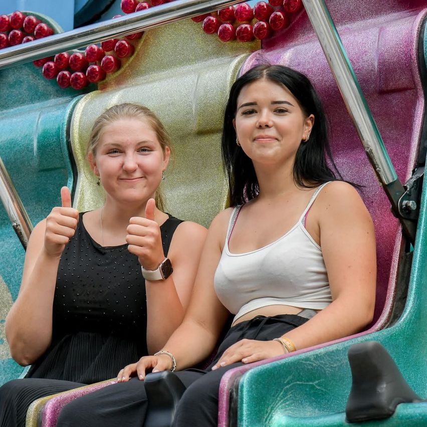 Zwei Frauen sitzen auf einer Achterbahn in einem Vergnügungspark. Eine zeigt Daumen hoch, und beide lächeln. Die Bahn hat bunte Dekorationen hinter ihnen.