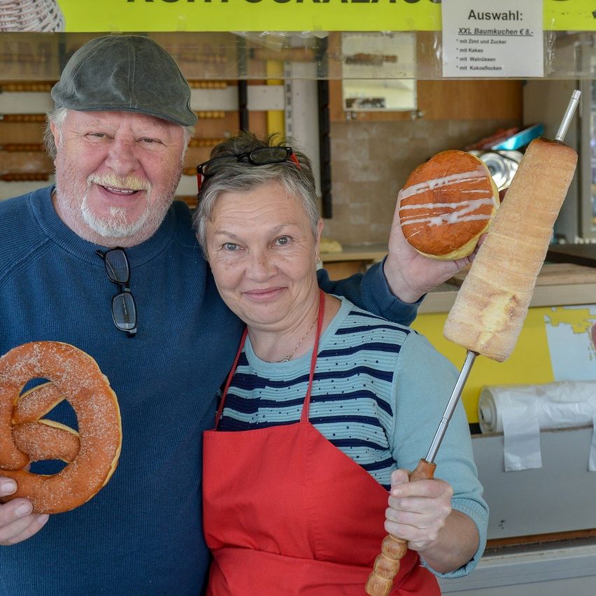 Ein Mann und eine Frau stehen vor einem Bäckereitresen, lächeln und halten Brezeln und einen Gebäckstab. Die Frau trägt ein rotes Schürzen. Hinter ihnen ist ein Menübrett und eine Glasvitrine.