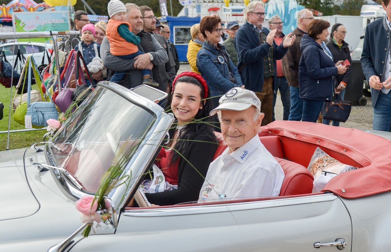 Ein älterer Mann und eine junge Frau sitzen in einem weißen Cabrio, umgeben von einer Menschenmenge auf einem Festival.