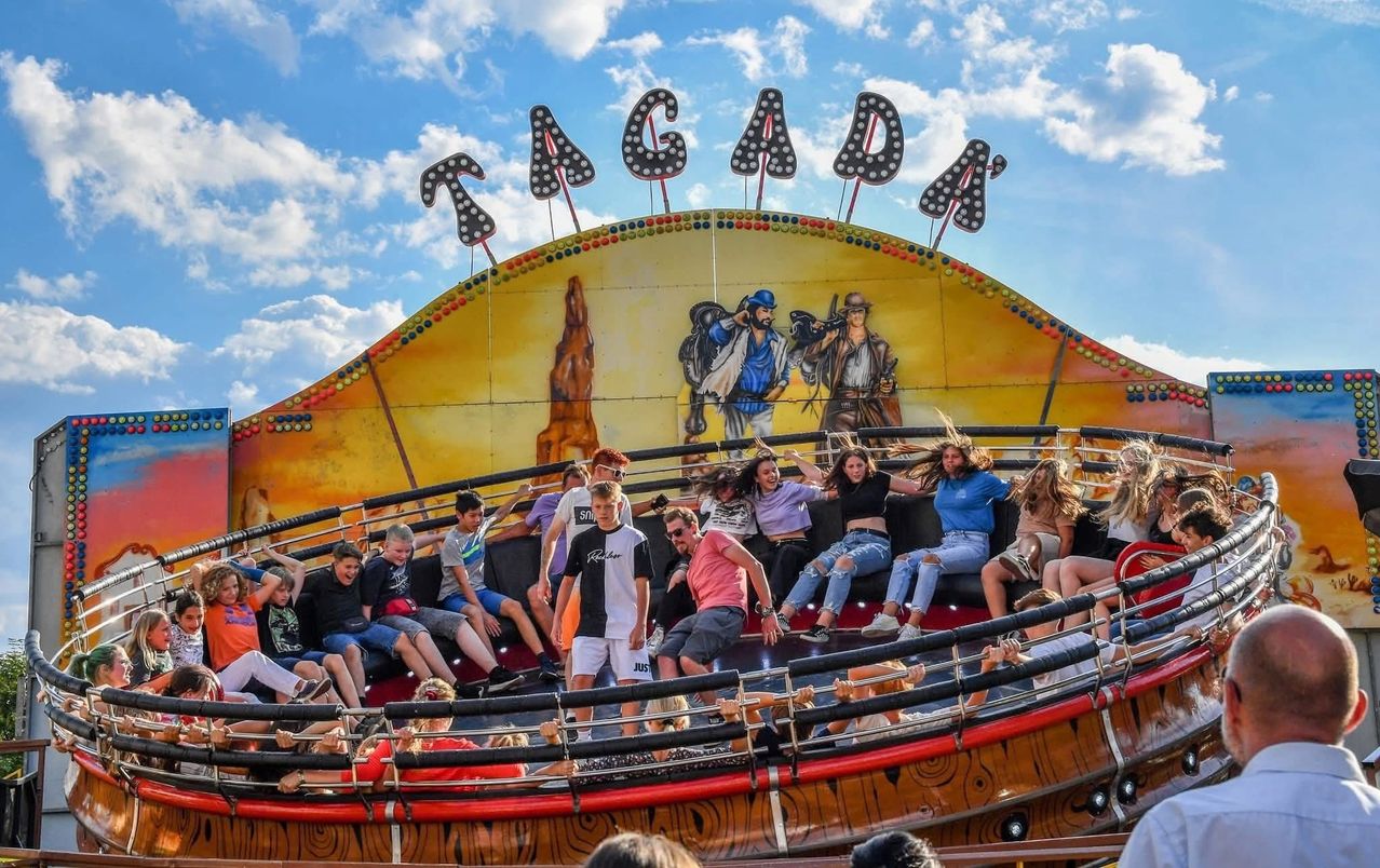Eine Gruppe von Menschen fährt auf einer rotierenden Jahrmarktattraktion namens Tagada, die mit Cowboys bemalt ist und sich vor einem blauen Himmel befindet.
