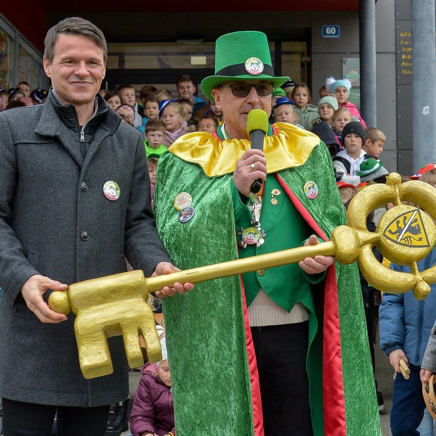 Zwei Männer stehen vor einer Menge; einer hält einen goldenen Schlüssel, der andere ein Mikrofon. Der Mann im grünen Kostüm trägt einen Hut mit Logo. Dahinter beobachten Kinder und Erwachsene.