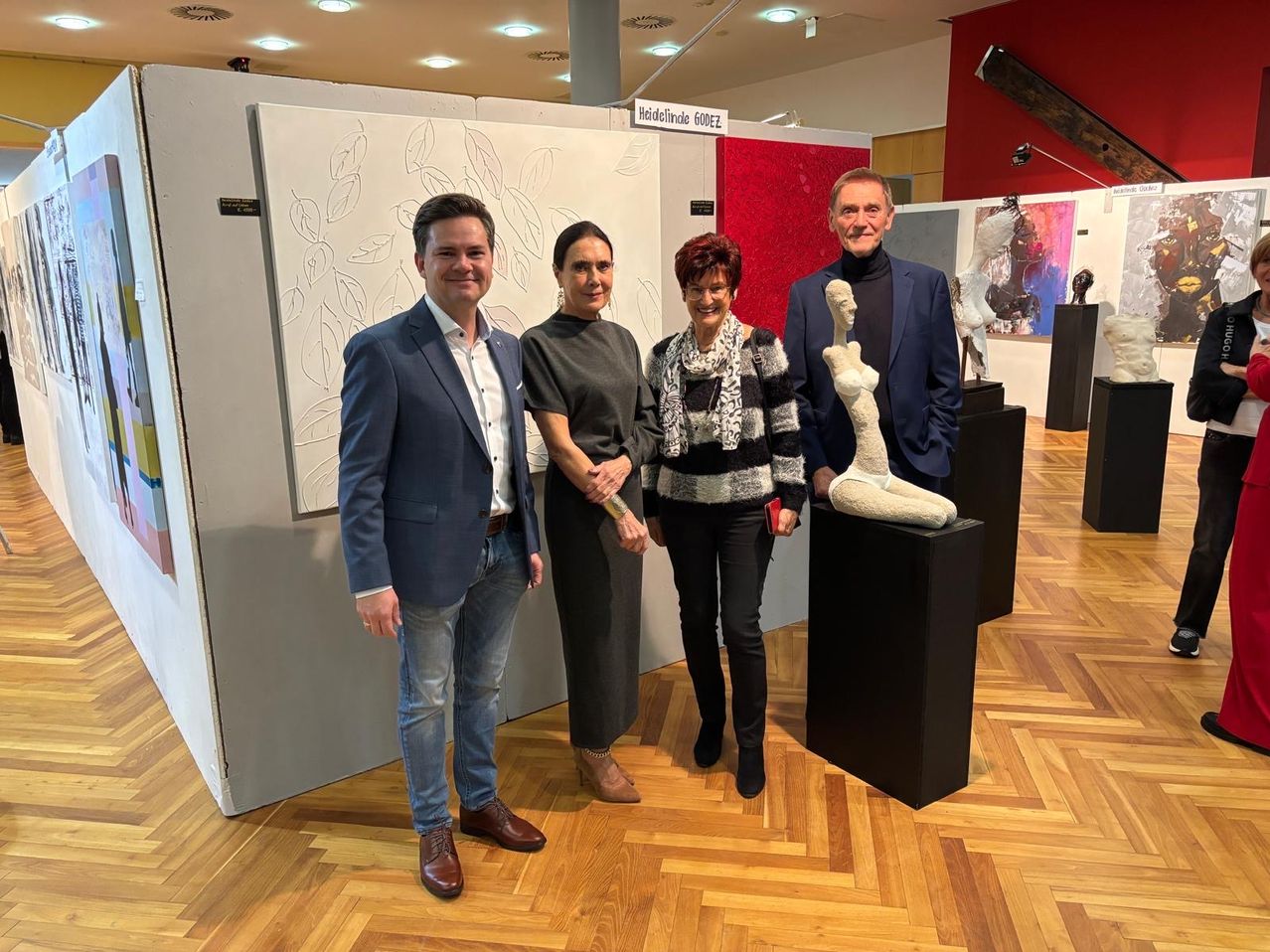 Vier Personen stehen vor einer Ausstellung in einer Kunstgalerie. Drei lächeln, und die Frau auf der rechten Seite hält ein rotes Objekt. Hinter ihnen befinden sich Skulpturen und Gemälde an den Wänden.