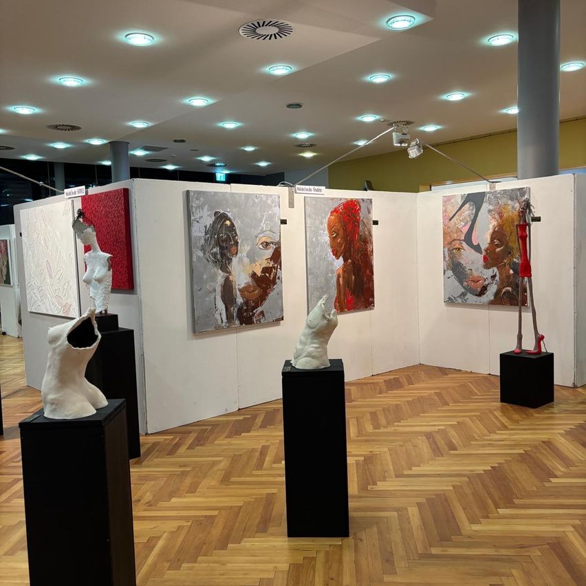 Eine Kunstgalerie mit Holzböden, weißen Wänden und schwarzen Podesten. Skulpturen und Gemälde sind ausgestellt, wobei ein Stück beleuchtet wird.