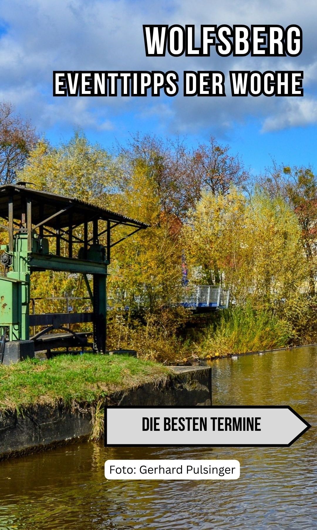 A green machine with a roof stands beside a river with a metal bridge in the background. Trees with autumn leaves surround the area. The sky is blue with some clouds.