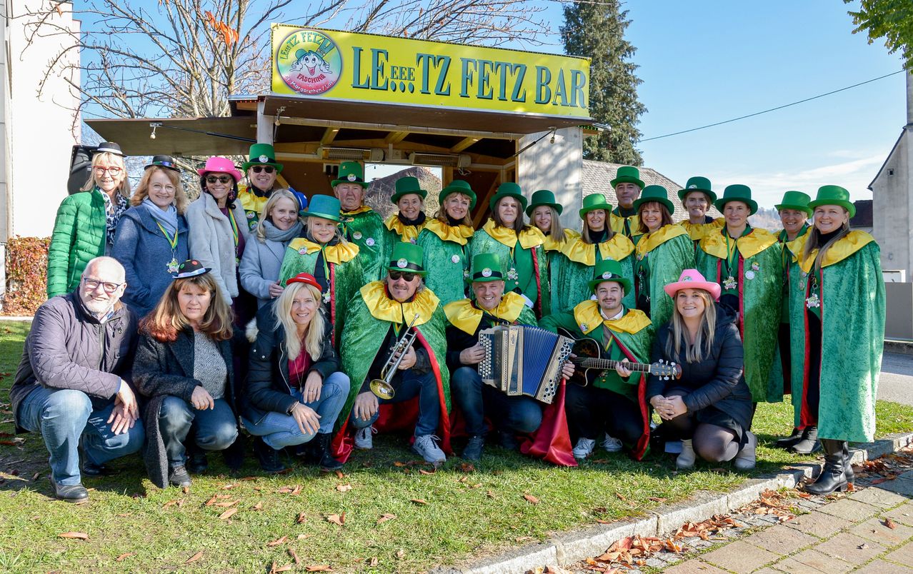 A group of people dressed in green and yellow costumes pose in front of a bar named Le...Tz Fetz Bar. Some of them are holding musical instruments.