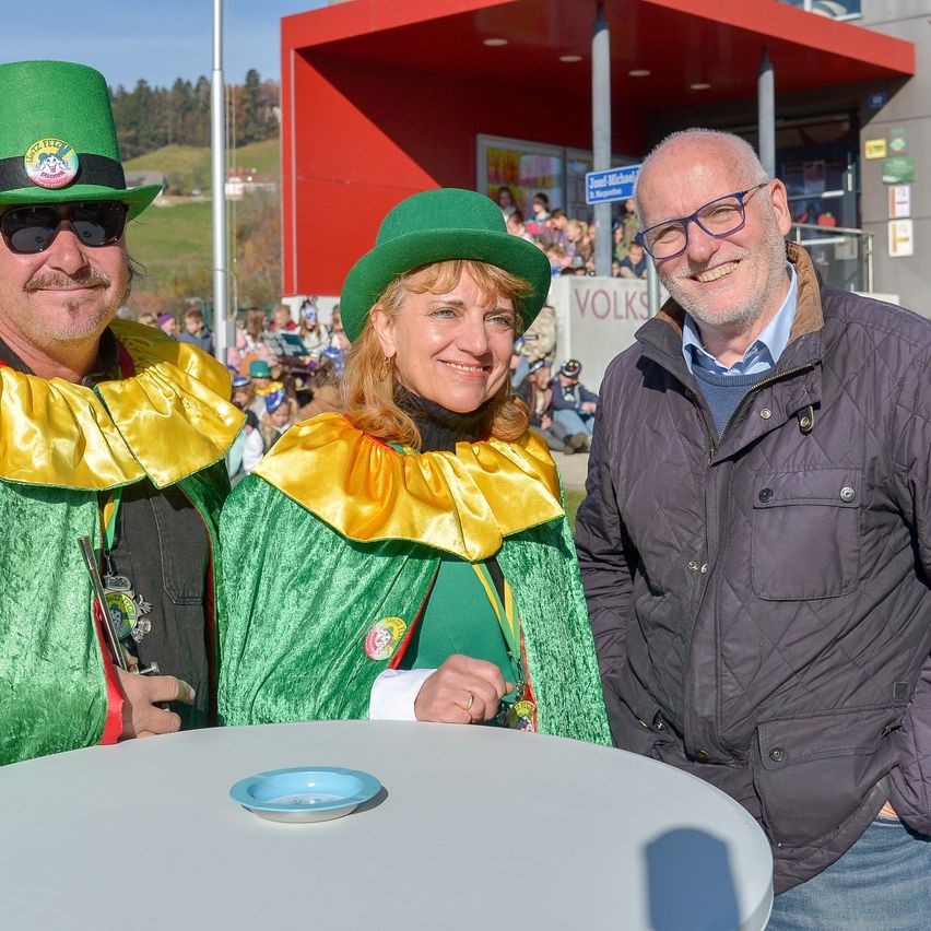 Three people are standing next to a table. Two are in festive costumes, one with a trumpet, and one in a jacket. Behind them is a crowd and a building.