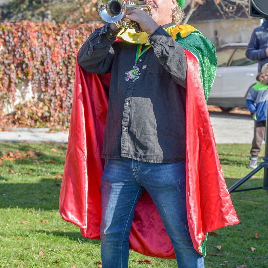 A man in a green, yellow, and red cape plays a trumpet on a grassy field. A car and a child are in the background.