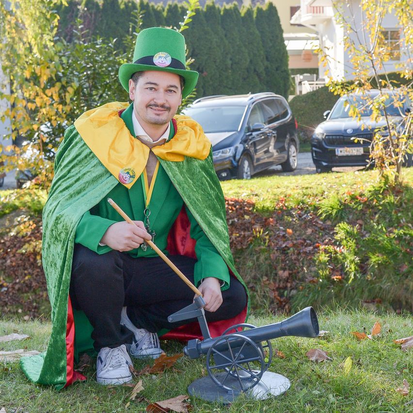 A man dressed as a magician in a green cape and hat holds a wand. He is kneeling next to a small cannon on a grassy lawn. Behind him are bushes, cars, and a building.