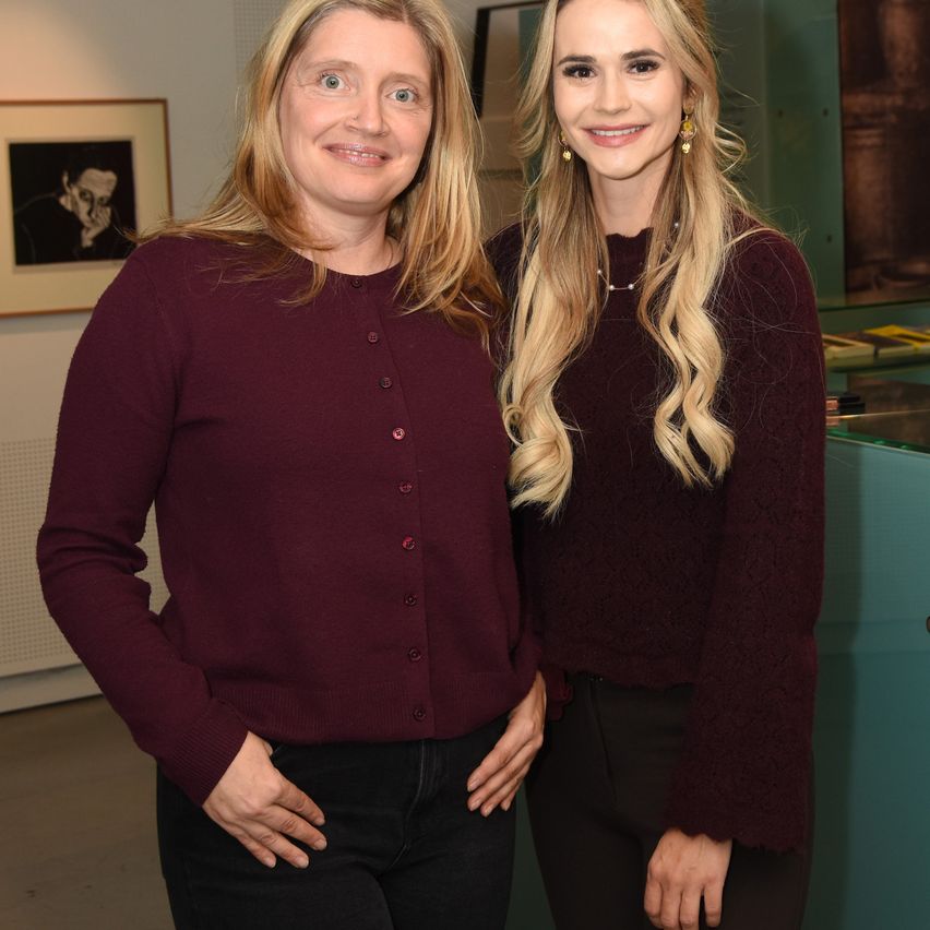 Two women in maroon sweaters and black pants smile for a photo in an indoor setting. The woman on the left has blonde hair, while the other has long blonde hair. A picture frame hangs on the wall behind them.