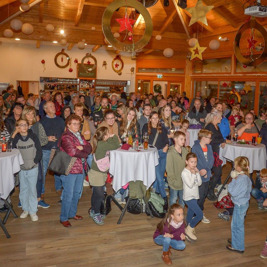 A crowded room with wooden flooring and tables covered in white cloths. Several people are gathered, some standing, some sitting. Decorations like stars and lanterns are hanging from the ceiling.