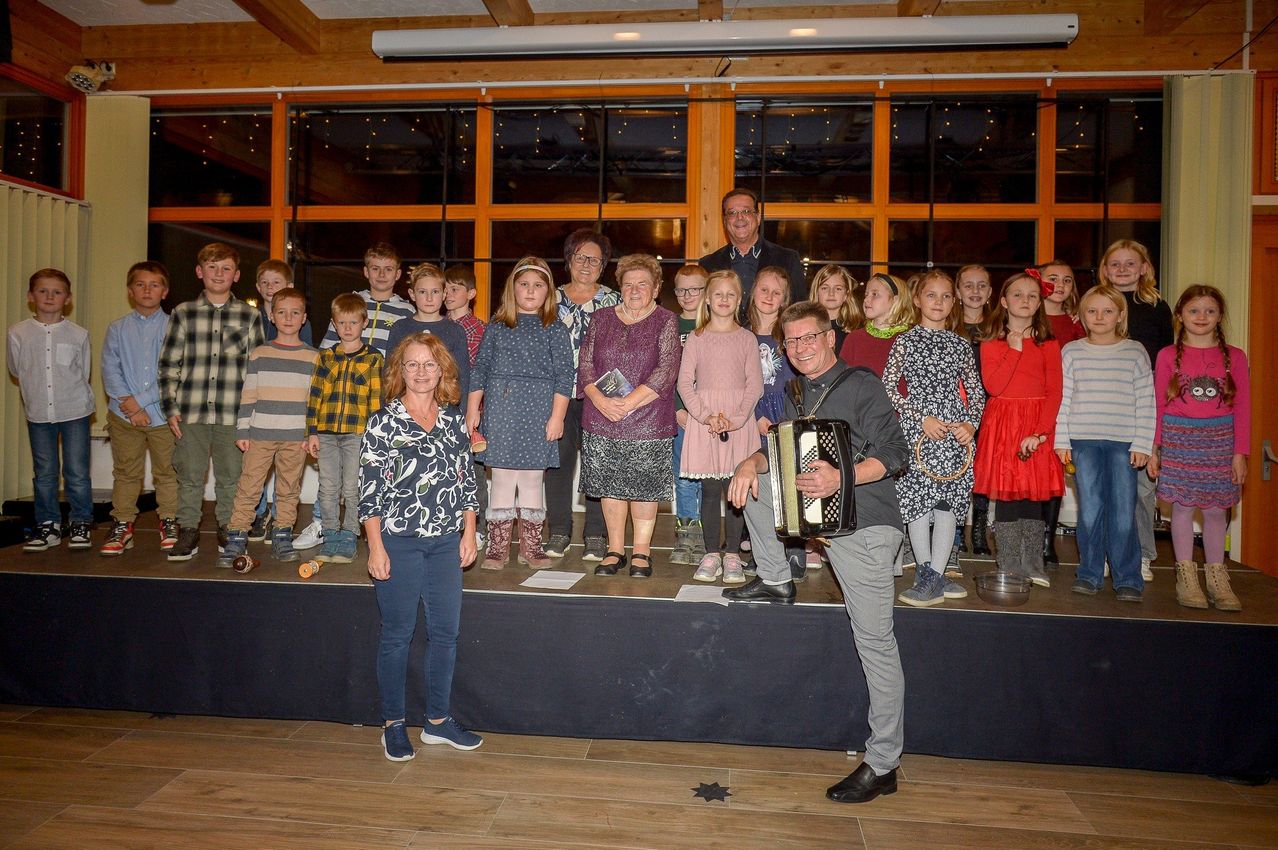 A group of children and adults stand on a stage with a man playing an accordion in front of them. They are dressed in various winter clothing.