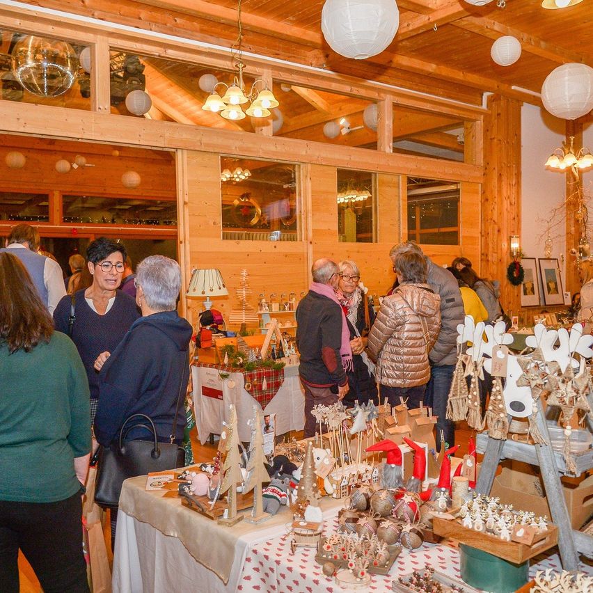 Several people stand in a wooden room, discussing around tables with various holiday items. The room is decorated with hanging lamps and Christmas decorations.