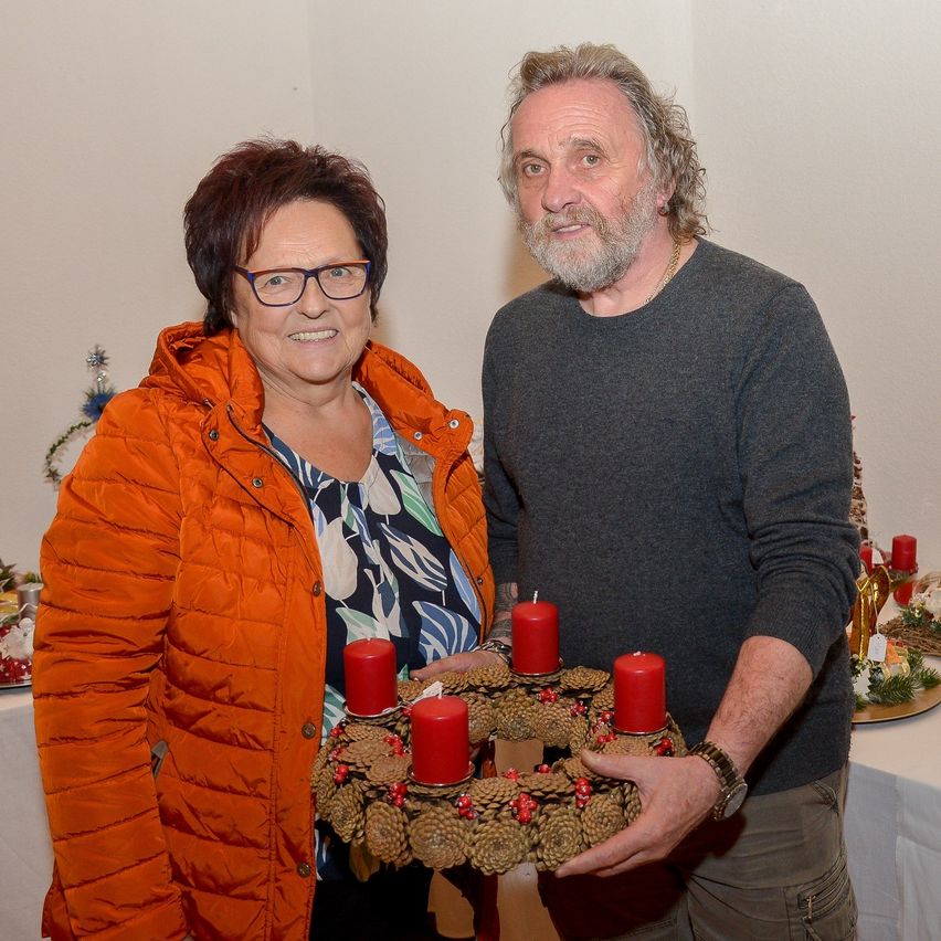 An elderly couple poses for a picture in a festive room with Christmas decorations. The woman wears an orange jacket, and the man wears a gray long-sleeve shirt. They hold a wreath with red candles.