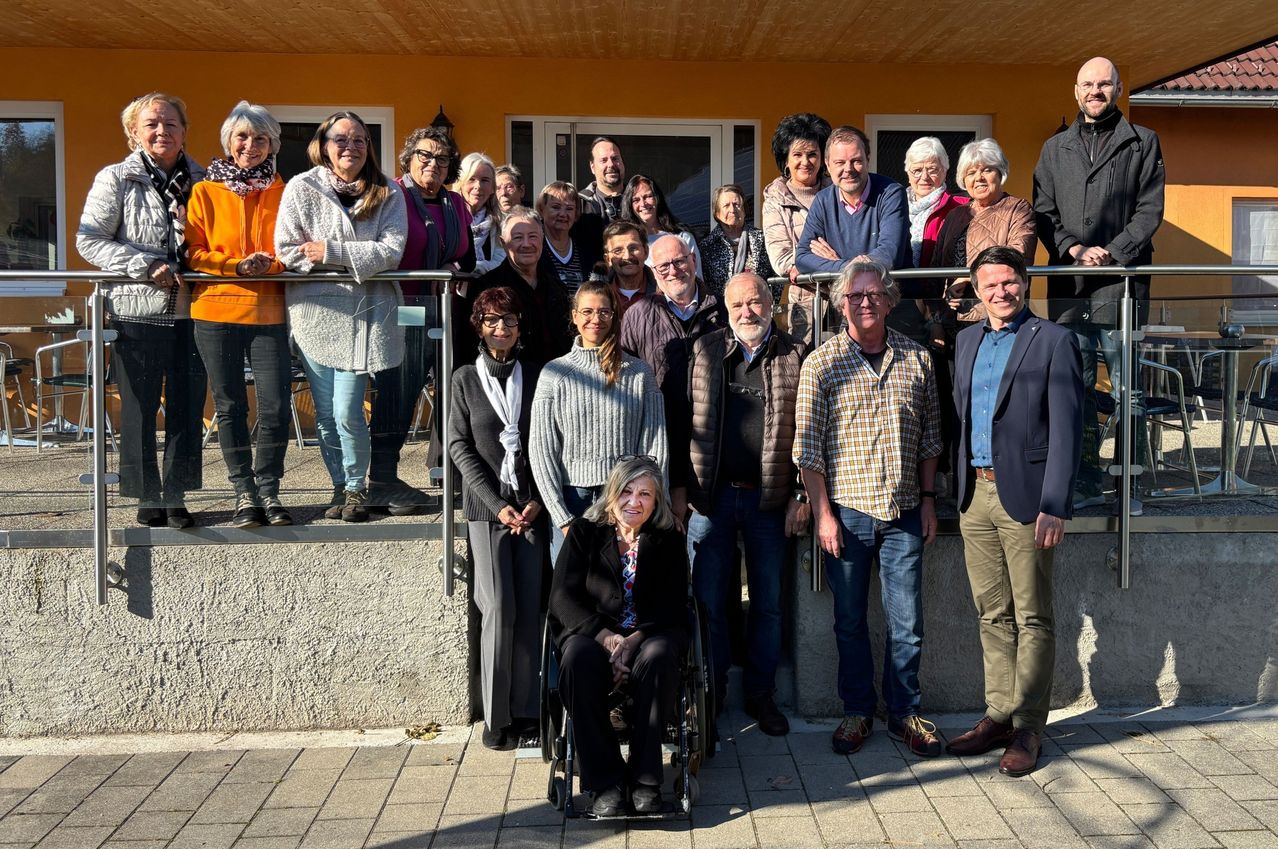 A diverse group of people stand in front of a yellow building, smiling for a group photo. A woman in a wheelchair is in the center.