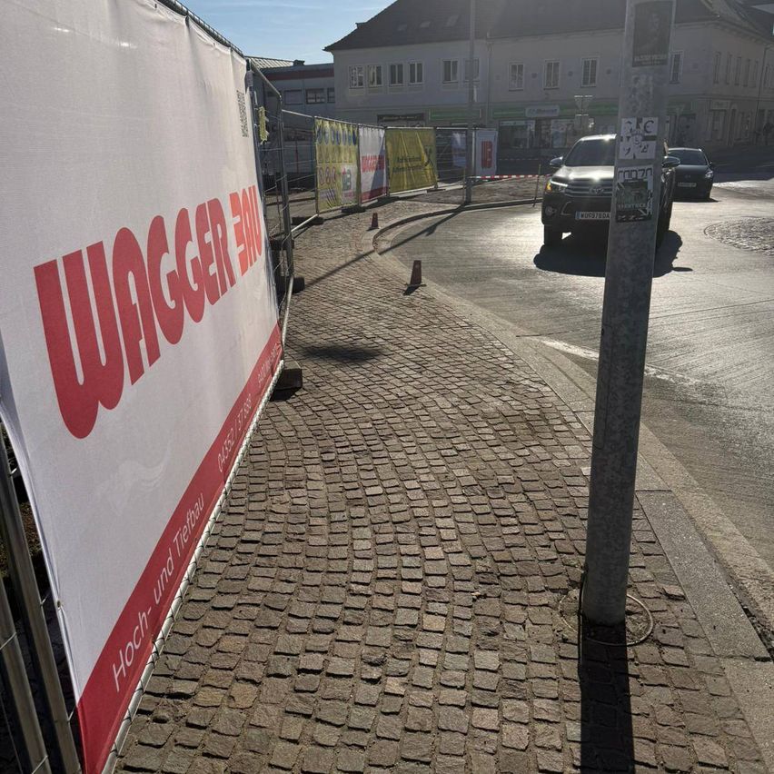 A red and white sign for Wagger Bau is placed on a brick sidewalk with a metal fence. Nearby, two cars are driving on the street.