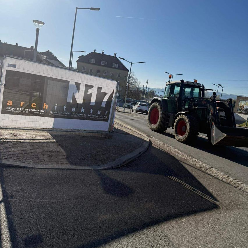 A tractor is driving on a road with a building in the background. An advertisement for architecture is visible on the side of the road.