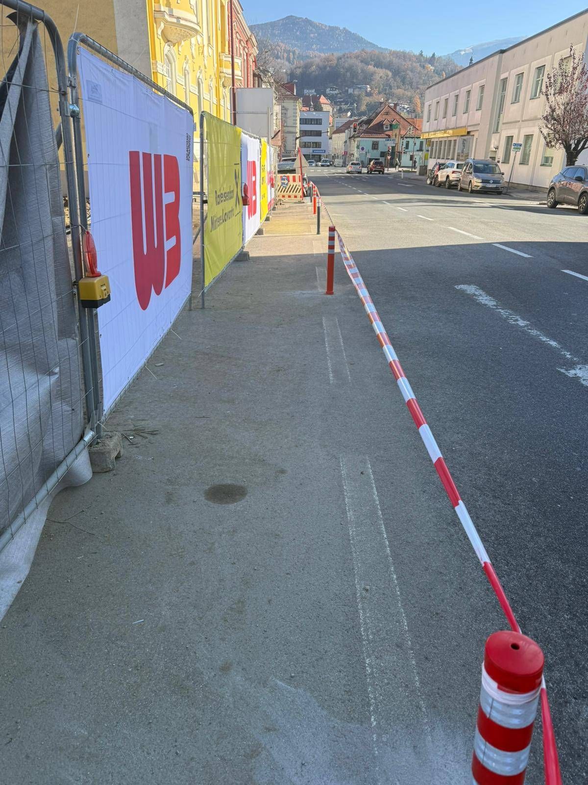 An urban street with a sidewalk barrier, advertisement banners, and parked cars. Buildings and traffic signs are visible.