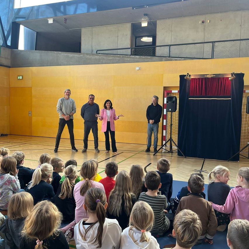Inside a gymnasium, three adults stand in front of a stage with a blue curtain. A crowd of children sit on the floor watching. The adults are gesturing and talking.
