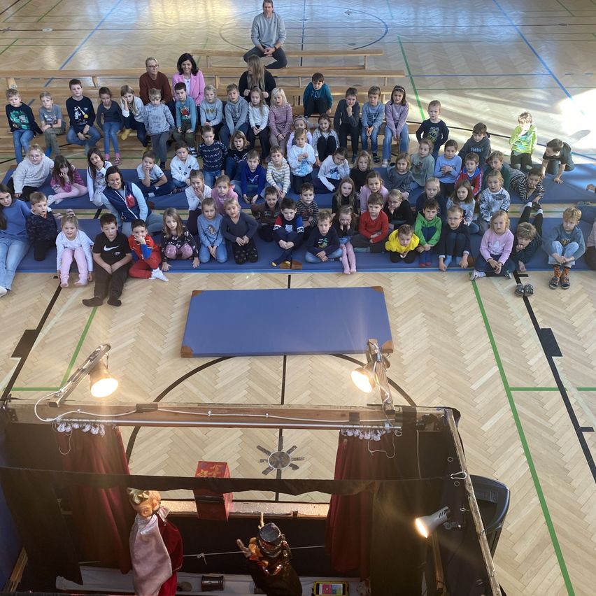 A group of children and adults gathered in a gym for a puppet show. Blue mats and lights are set up on the floor.