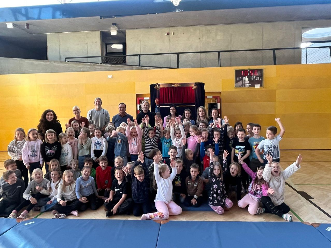 A group of children and adults are posing for a photo in a gymnasium. The children are sitting and standing, some raising their hands. Adults are positioned at the back, with a scoreboard and a stage visible.